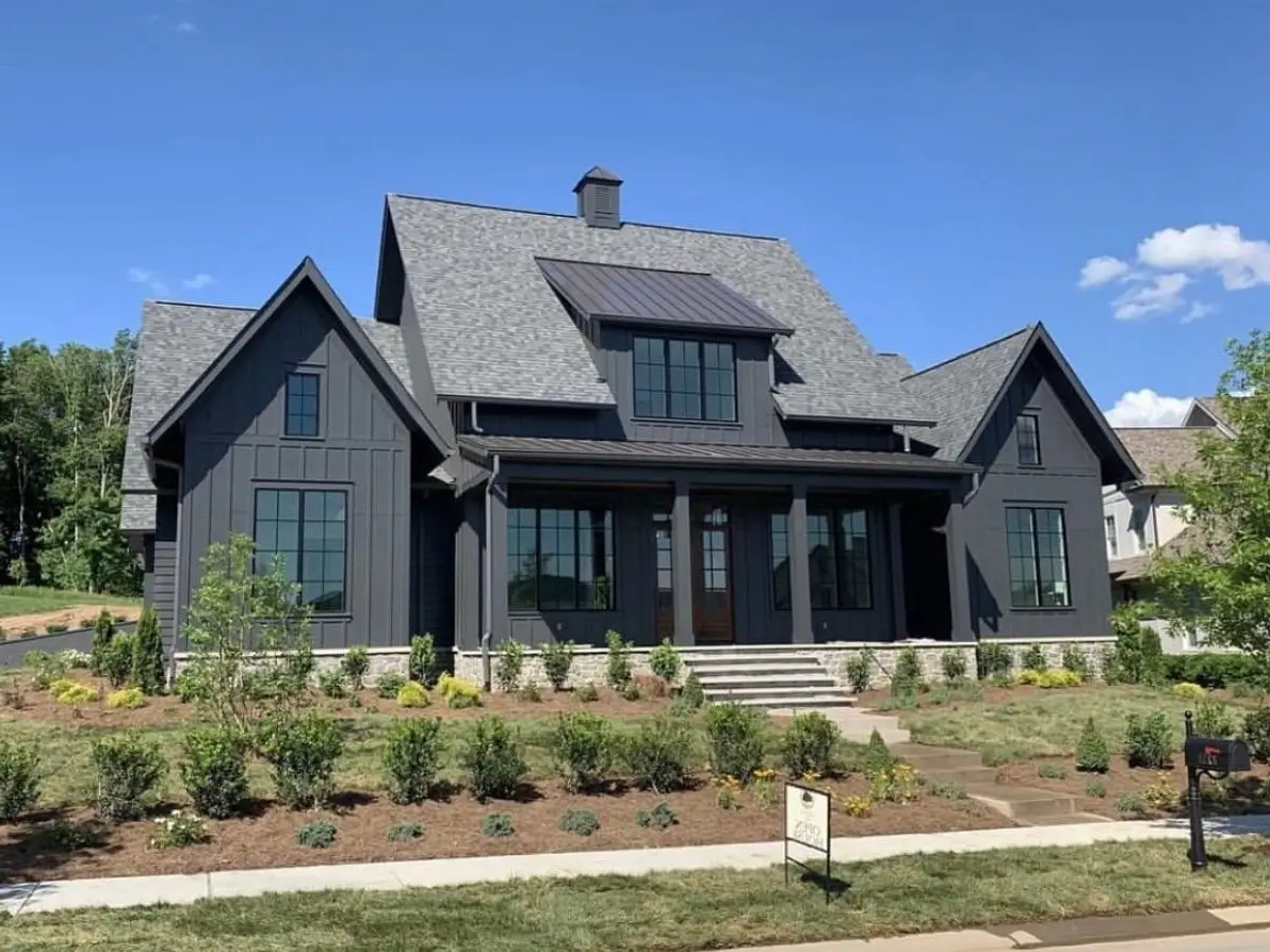 A modern black farmhouse exterior with striking layered gables, a dark metal roof accent, and a light stone foundation, showcasing excellent curb appeal.