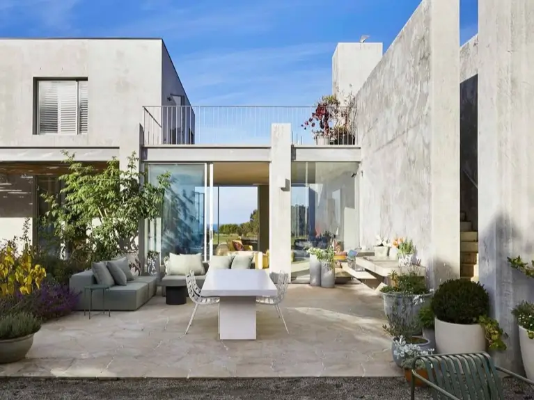 The sunlit patio of a modern brutalist house, featuring a comfortable outdoor sofa, dining area, and large glass doors that connect to the interior living space.