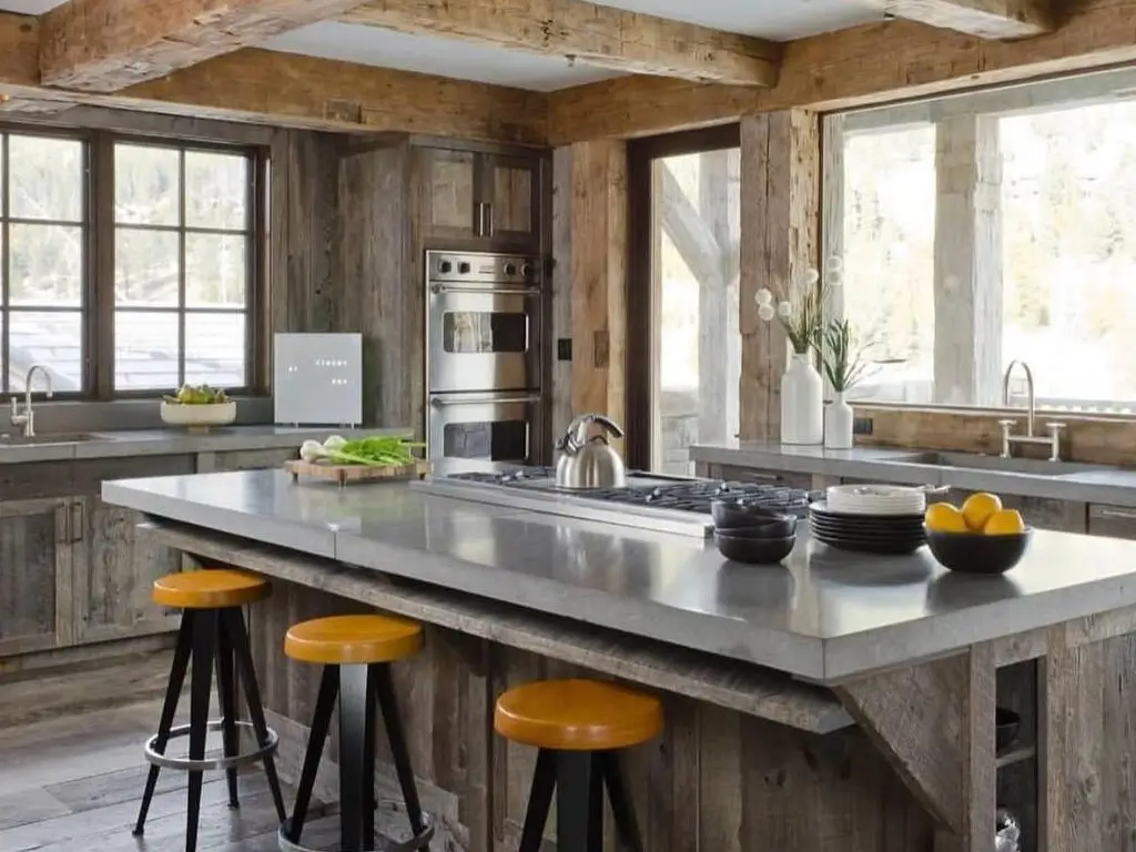 A modern rustic kitchen featuring weathered wood cabinets, a large concrete island with seating, and exposed wooden ceiling beams.