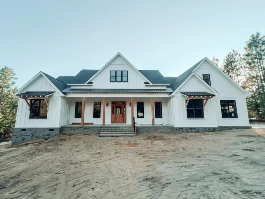 A newly constructed modern white farmhouse exterior with vertical siding, a dark roof, black windows, and a central wooden front door. The home features a brick foundation and is set on a dirt lot with pine trees.