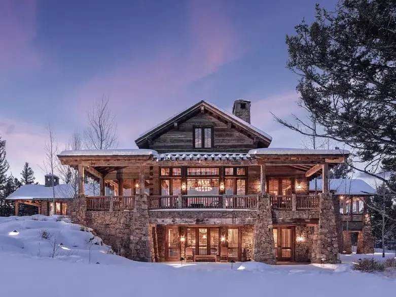 16 Mountain Mansions That Define Luxury Alpine Living 3 A rustic log and stone mountain mansion covered in snow at dusk, with warm light glowing from the windows, creating a cozy sanctuary.