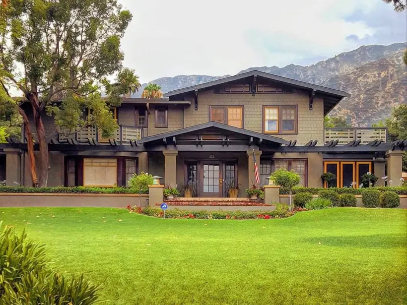 A stately craftsman house exterior in earthy tones, featuring a wide porch and prominent pergolas with a mountain landscape behind it.