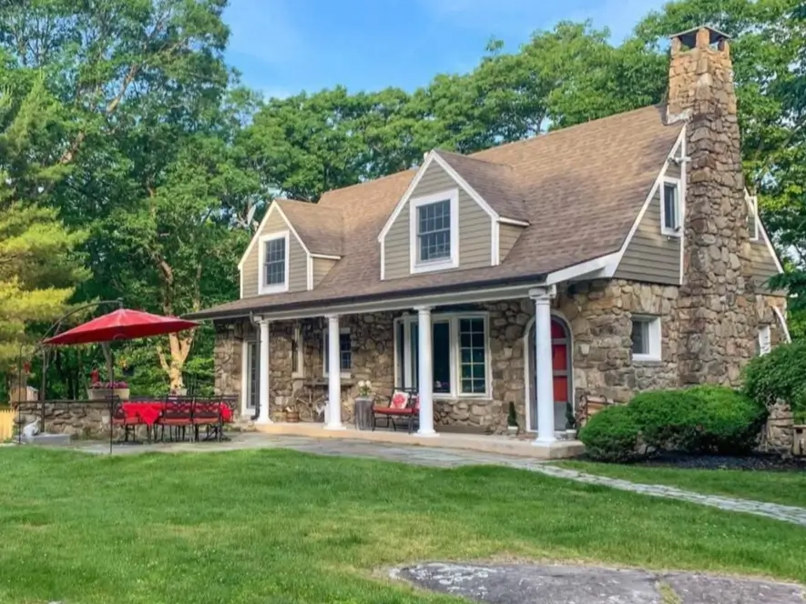 A charming stone cottage house with a large fieldstone chimney and a covered front porch, set in a lush green landscape.