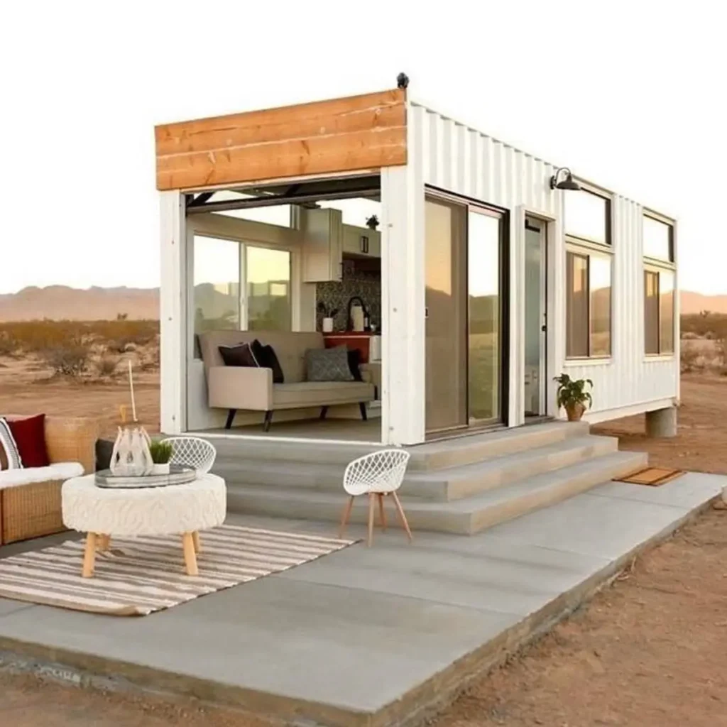 A white small shipping container home in a desert setting, featuring a large glass wall that opens onto a modern concrete patio with stylish outdoor furniture.