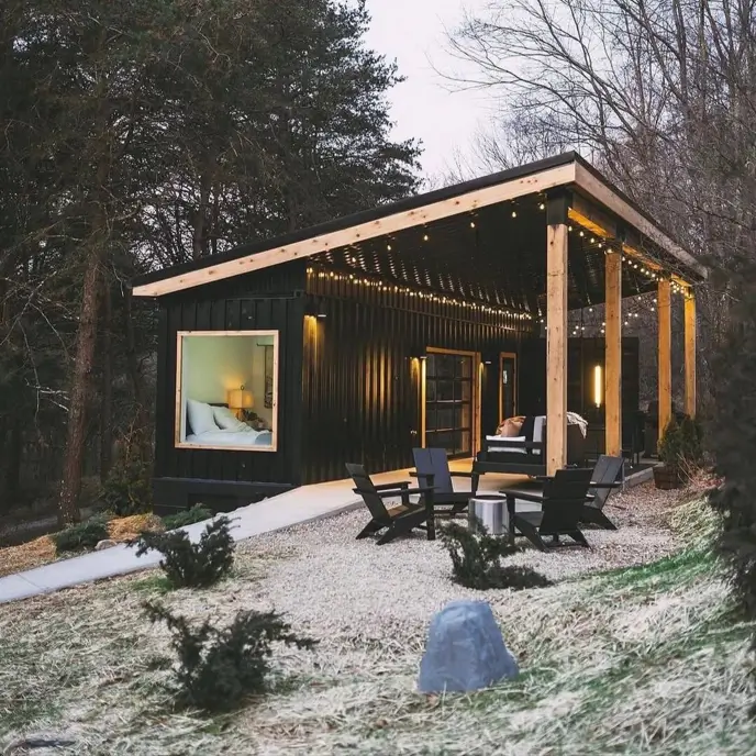 A modern black tiny container house design in the woods with a large covered porch, string lights, and a gravel patio with chairs.