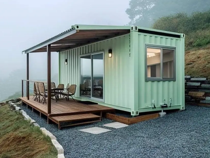 A light green small shipping container home on a foggy hillside, featuring a covered wooden porch with an outdoor dining set.