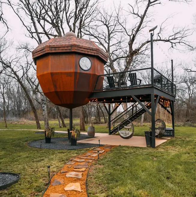 A unique house design of an acorn-shaped treehouse made of rusted metal with an attached deck.