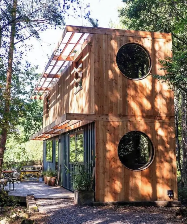 A two-story small container house design with wood cladding and large circular windows, set in a forest.