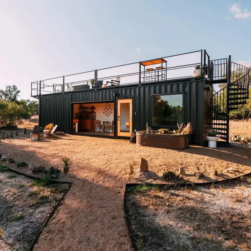 A black small shipping container home with a full rooftop deck accessed by a spiral staircase in a desert landscape.