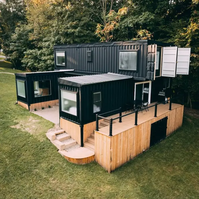 A unique house design made from six stacked black shipping containers of various sizes, with a large wooden deck nestled by trees.