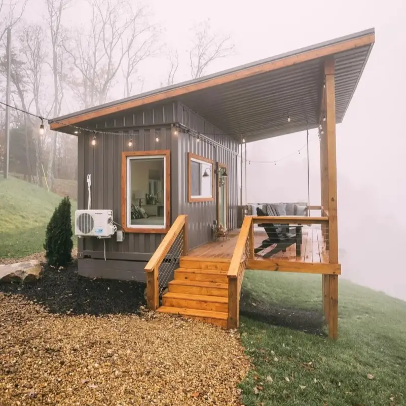 A dark gray small container home on a foggy hill, with a wooden elevated porch, a sloped roof, and string lights.