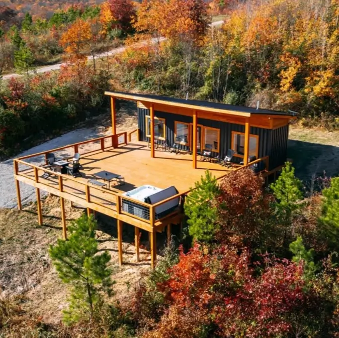 An aerial view of a black small container home with a large wraparound wooden deck and hot tub, surrounded by colorful autumn trees.