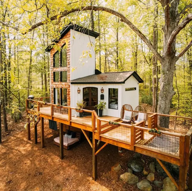 A unique house design nestled in a forest, featuring an elevated wooden deck and a mix of white siding and brick.