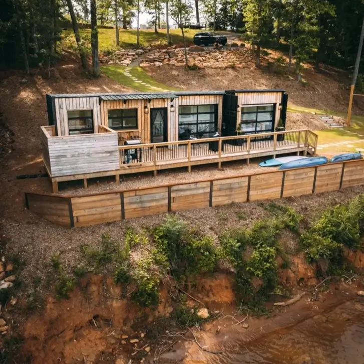 A long small shipping container home with wood accents built into a lakeside hill, featuring a long deck and large glass doors.