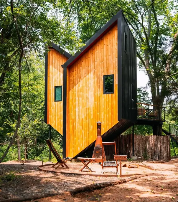 A unique house design of an angular wood and black cabin elevated in a forest clearing with a fire pit.