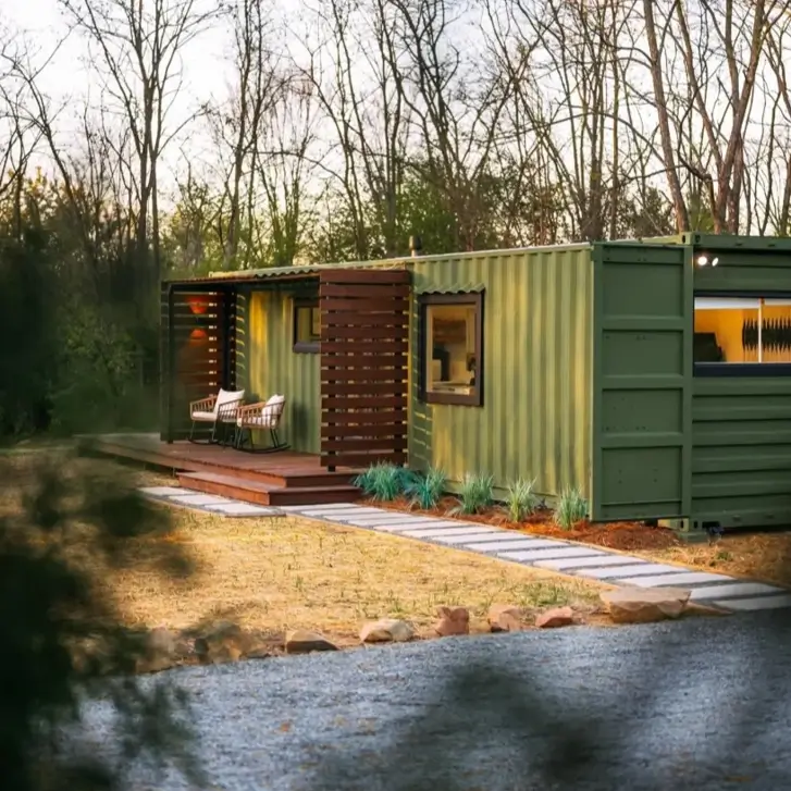 An olive green small shipping container home at dusk, featuring a modern wooden slat privacy screen on its deck.