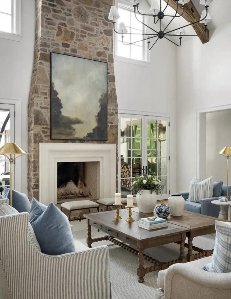 Modern farmhouse living room with a large stone fireplace, high ceilings with wood beams, and blue and white furniture.