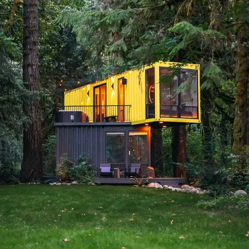 A two-story small container home in the woods, with a black lower level and a bright yellow upper level featuring a balcony and large windows.