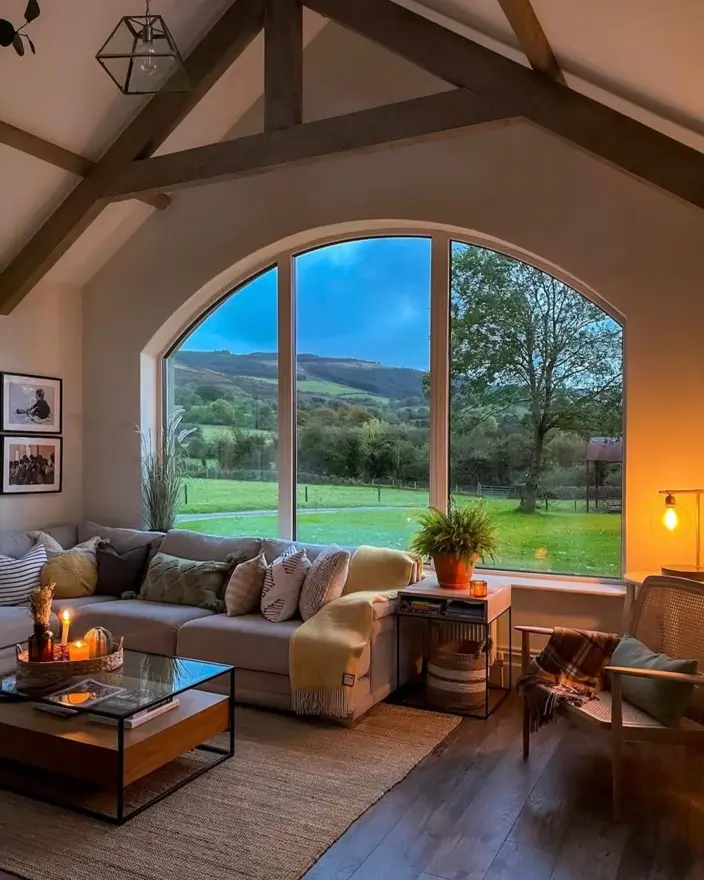 Modern farmhouse living room with vaulted ceiling, wood beams, and a large arched window overlooking a green landscape.