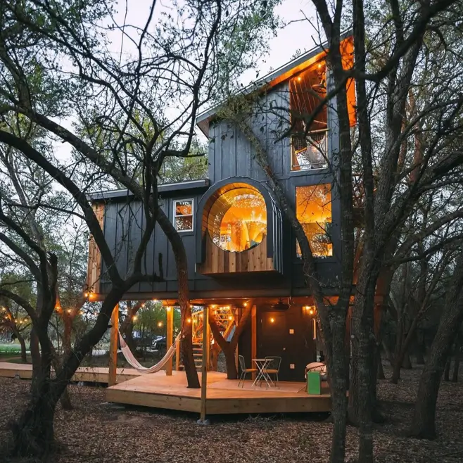 A unique house design of a modern dark treehouse with a large arched window glowing at dusk.