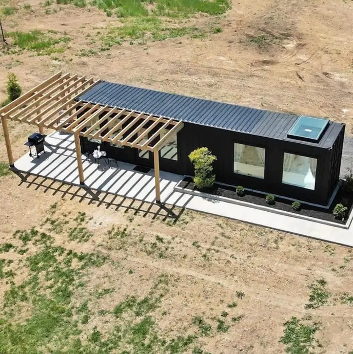 An aerial view of a sleek black small container house featuring a modern wooden pergola over a concrete patio and a large skylight.