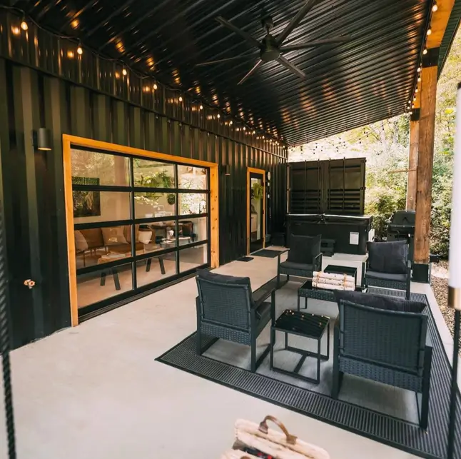 A modern black small shipping container home at dusk with a large glass garage-style door opening onto a covered patio with stylish outdoor furniture.