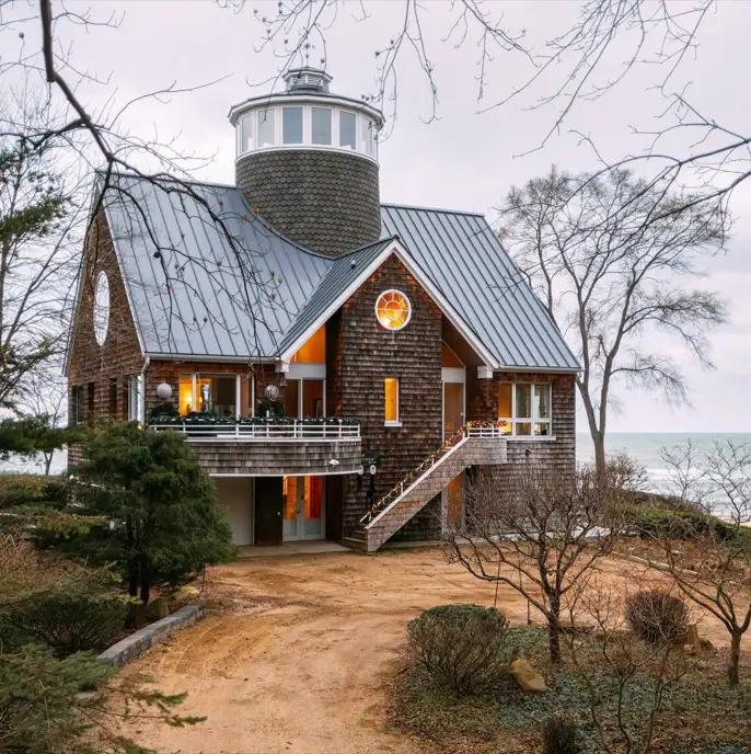 A unique house design with a lighthouse turret, cedar shake siding, and a metal roof on a lakeshore.