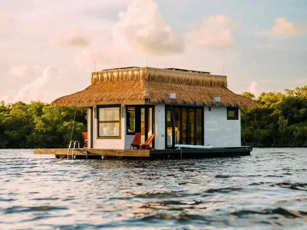 A unique house design featuring a modern floating home with a large thatched roof on the water at sunset.