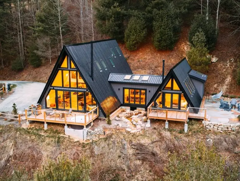 A stunning house design of a modern compound with two black A-frame cabins connected in a wooded landscape.
