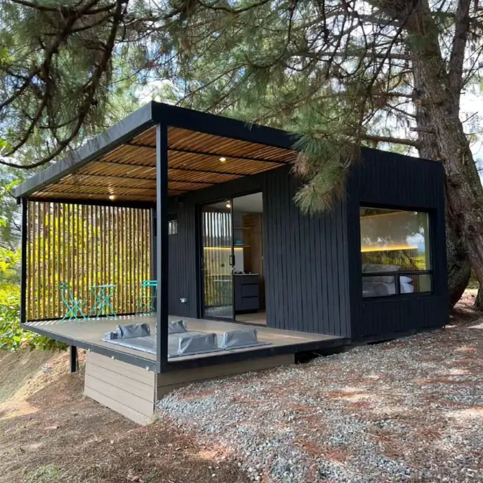 A modern black tiny container house design with a wooden deck, a built-in hot tub, and a slatted privacy screen, nestled under trees.