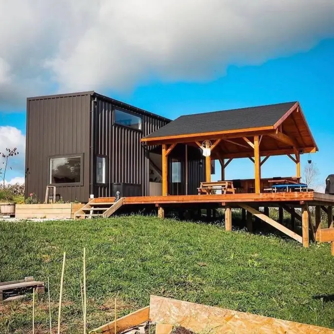 A dark brown small shipping container home on a hill, connected to a large elevated wooden deck with a covered gazebo area for outdoor entertaining.