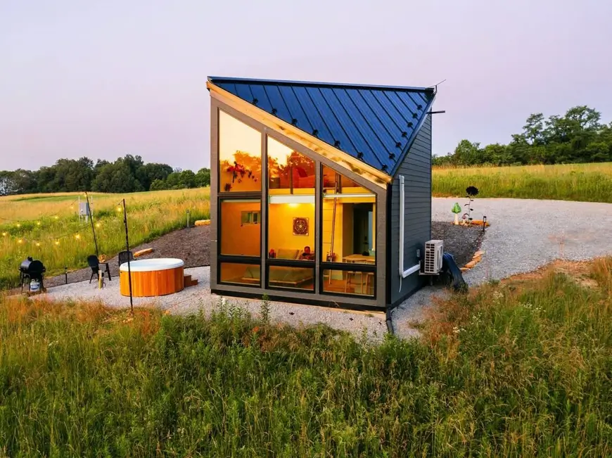 A beautiful modern house design of a compact cabin with an asymmetrical roof and glass wall, beside a hot tub in an open field.