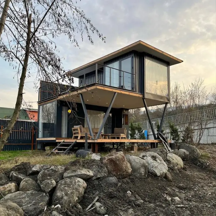 A two-story small shipping container home on angled stilts, with extensive glass walls and an upper balcony overlooking a wooden deck.