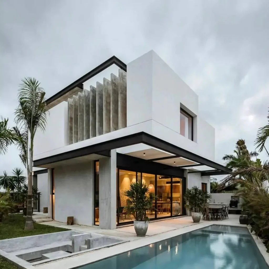 A beautiful modern house design with white and concrete walls, a black frame, and a poolside patio with tropical plants.