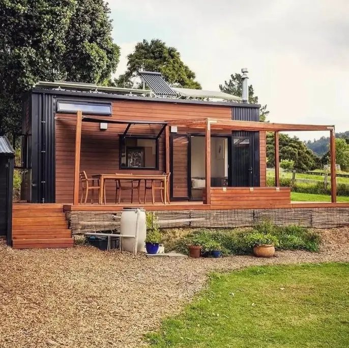 A small container home with natural wood cladding and a large multi-level deck that includes an outdoor dining area under a wooden pergola.