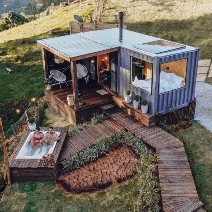 Aerial view of a gray tiny container house design on a hill, with a large bedroom window, wooden walkway, and a separate hot tub deck.