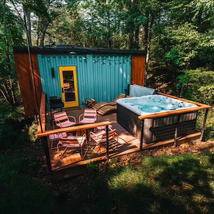 A teal small shipping container home in the woods with a yellow door, featuring a wooden deck with a hot tub and outdoor seating.