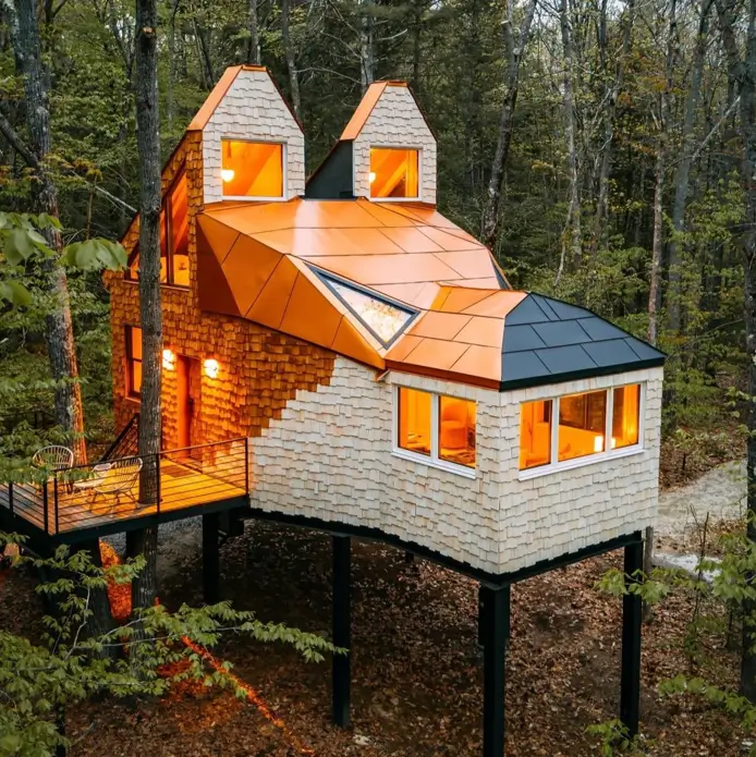 A unique house design of a treehouse in Maine shaped like a fox, with cedar shakes and a copper-colored roof, elevated in a forest.