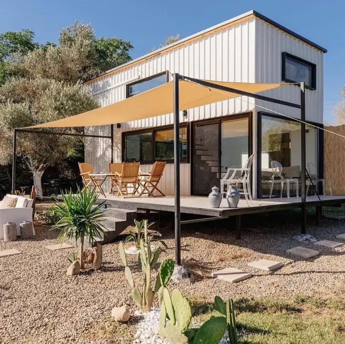 A white two-story small container home with a large wooden deck shaded by a tan sun sail, surrounded by a gravel and cactus garden.