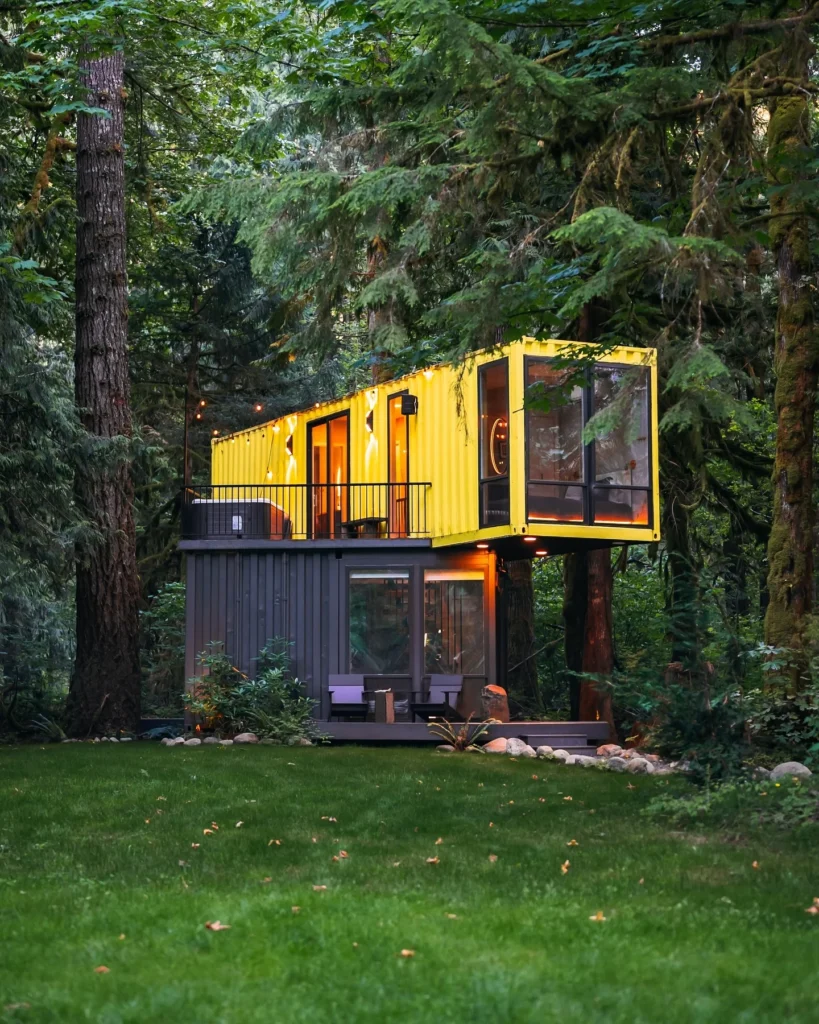  Bright yellow shipping container home elevated on stilts in a forest setting.