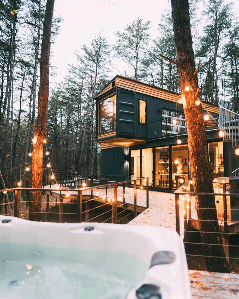 Multi-level shipping container home in a forest with a hot tub and large deck.