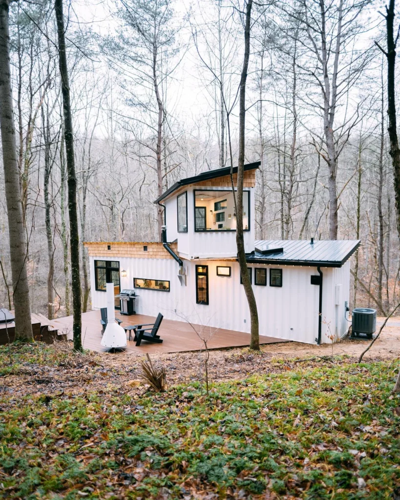 White two-story shipping container home with an angled roof in a wooded area.