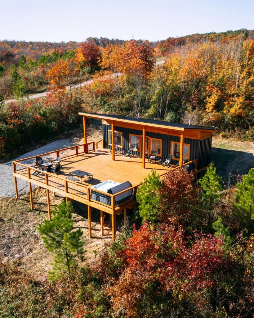 Small shipping container home with a large wooden deck and hot tub, overlooking a valley.