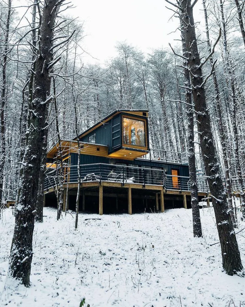 Shipping container home with angled roof and large deck in a snowy forest.