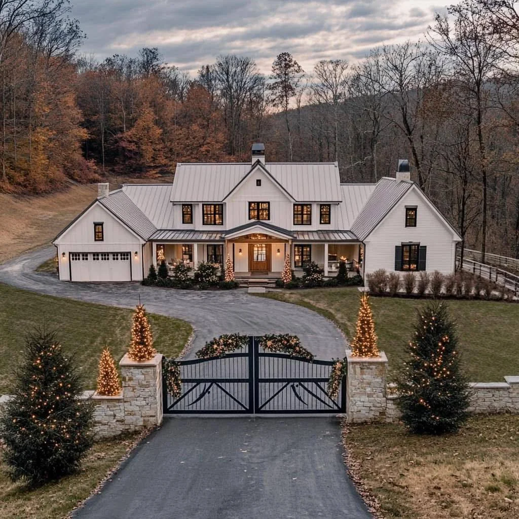 Modern farmhouse decorated for Christmas with lighted trees, gated entrance, stone pillars.