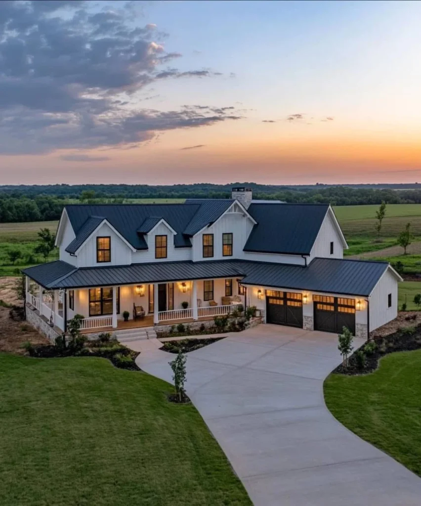 Modern farmhouse with wrap-around porch, black and white exterior, curved driveway, sunset view.