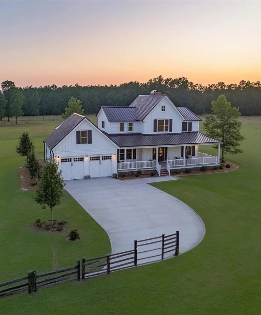 Two-story modern farmhouse with metal roof, wrap-around porch, white siding, split-rail fence.