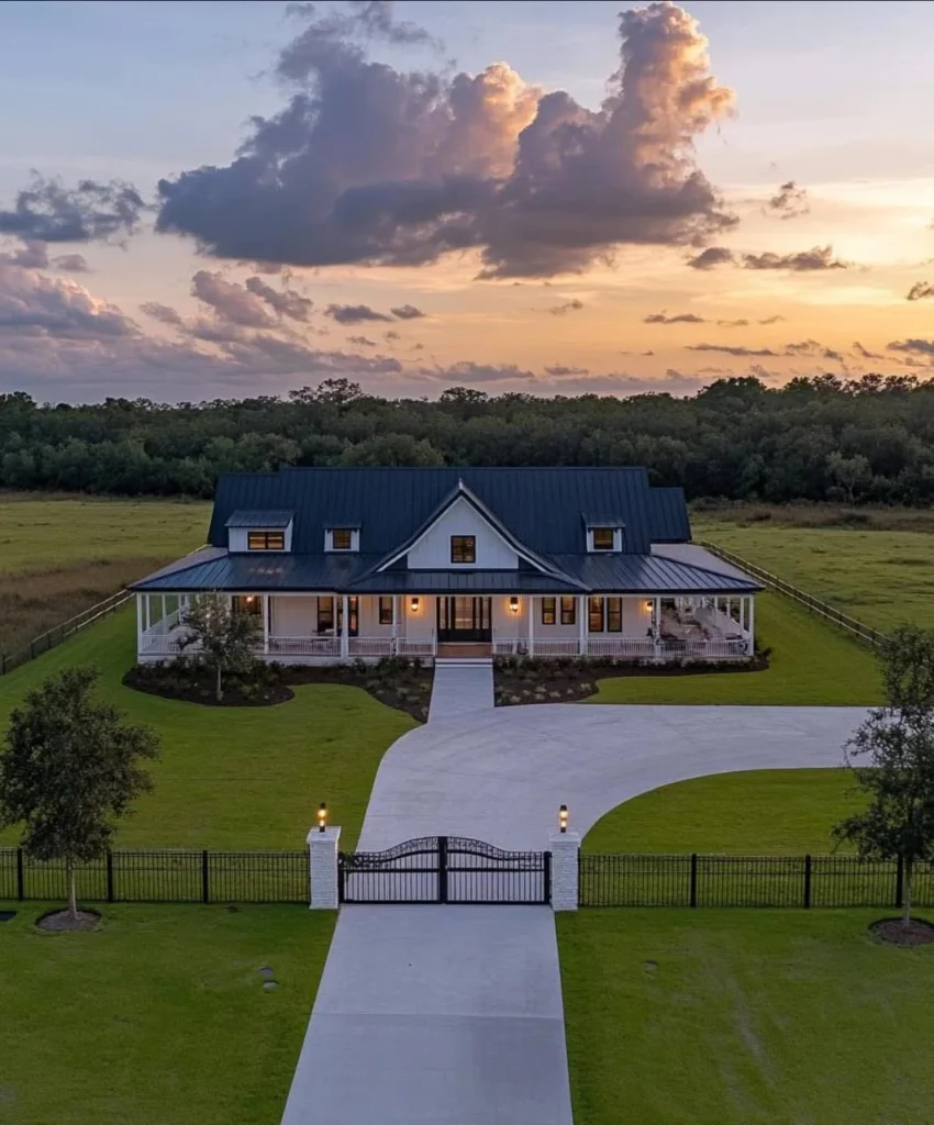 Modern farmhouse with wrap-around porch, metal roof, long driveway, gated entrance, large lawn.