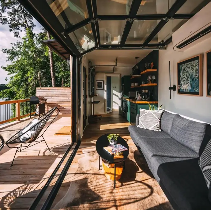 The interior of a tiny container house with a retractable glass wall fully open to a wooden deck, showing a seamless indoor-outdoor living space.