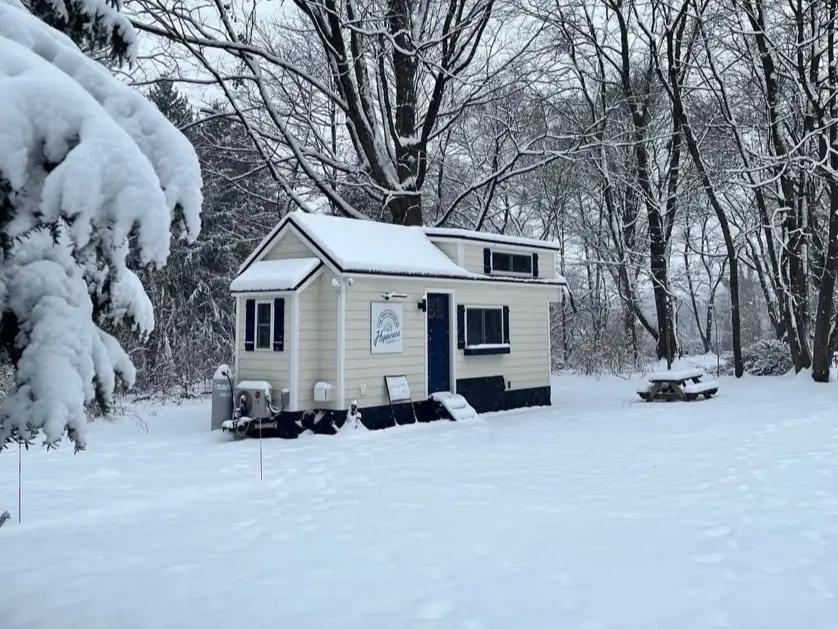 A cozy cream-colored tiny house nestled in a tranquil, snow-covered winter forest.